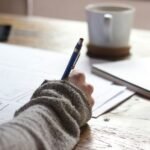 Main tenant un stylo bleu, écrivant sur des documents, avec un mug et un carnet à côté.