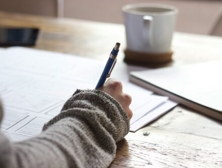 Main tenant un stylo bleu, écrivant sur des documents, avec un mug et un carnet à côté.