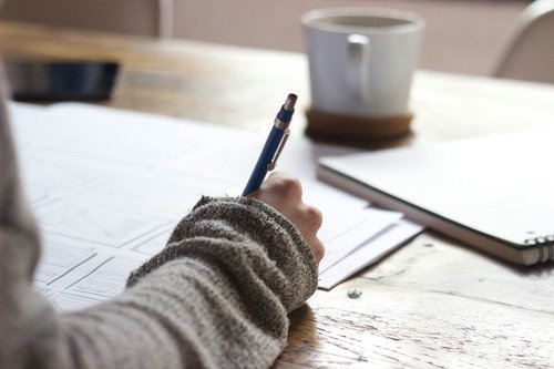 Risques psychosociaux : Prévention en entreprise Main tenant un stylo bleu, écrivant sur des documents, avec un mug et un carnet à côté.