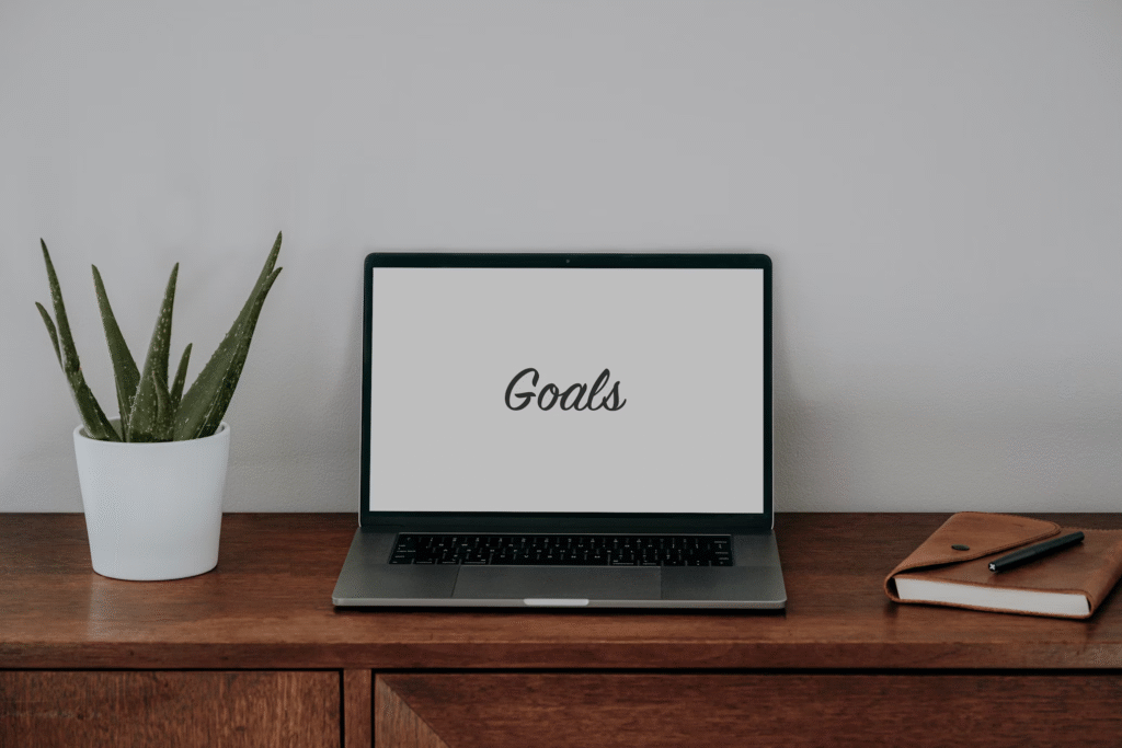 Bureau minimaliste avec plante, ordinateur affichant 'Goals' et carnet pour fixer des objectifs.